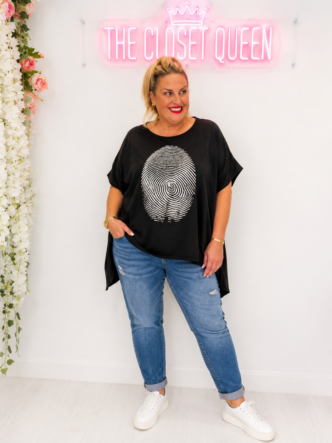 A woman modeling a black short-sleeve t-shirt with a silver glittery spiral fingerprint graphic on the front, paired with blue jeans and white sneakers, standing in front of a white wall with a pink neon sign that reads 'THE CLOSET QUEEN'.