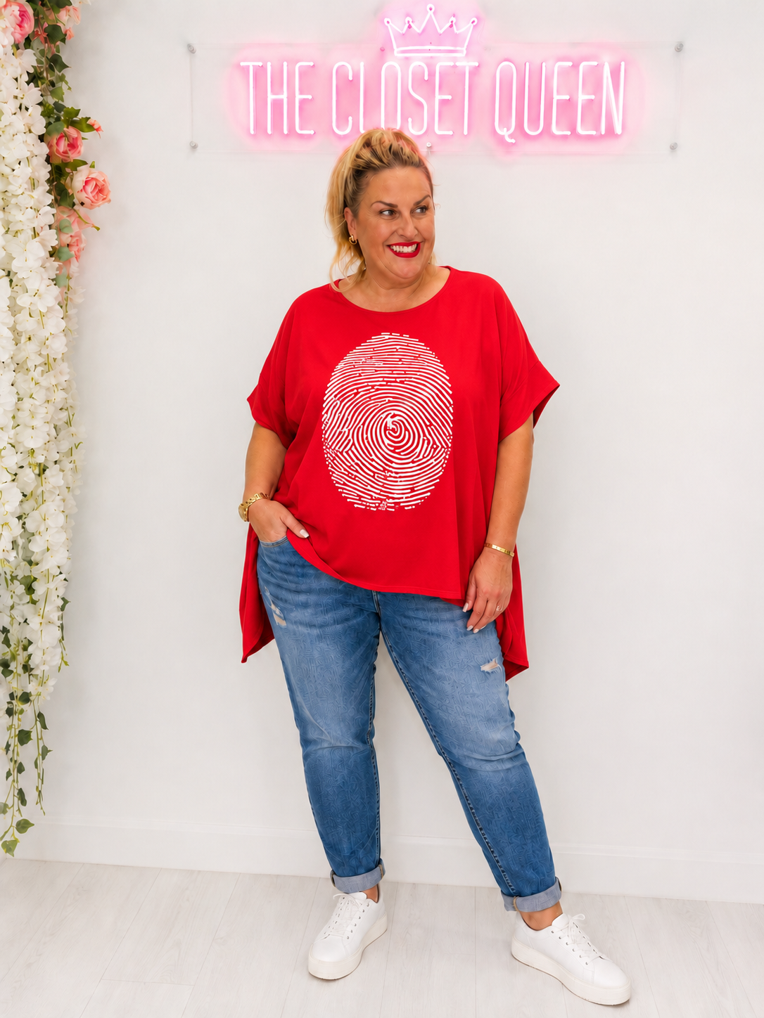 A woman modeling a bright red oversized t-shirt with a white circular fingerprint graphic on the front, paired with blue jeans and white sneakers, standing in front of a white wall with a pink neon sign that reads 'THE CLOSET QUEEN'.
