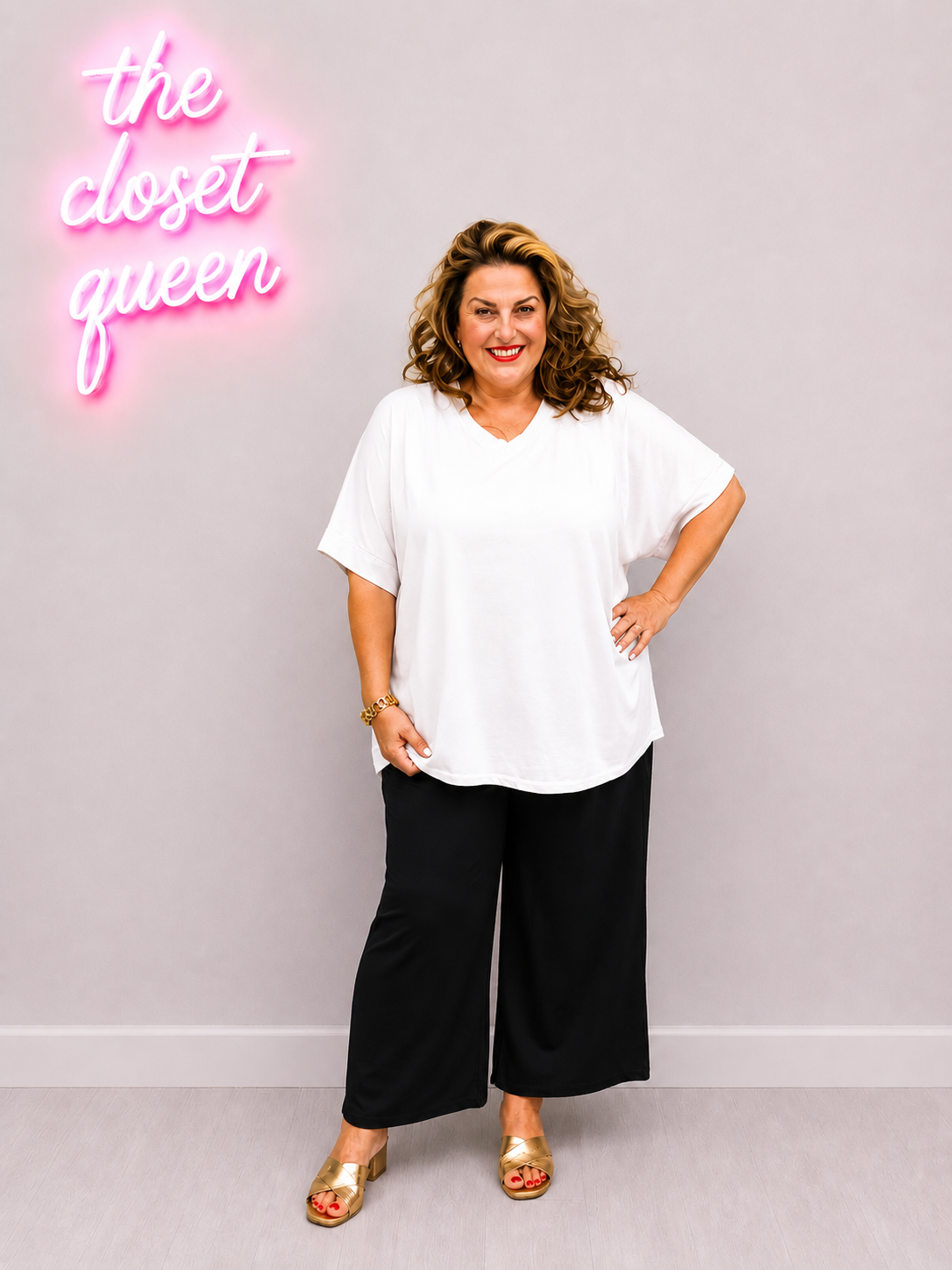 Woman in a white top and black pants standing in front of a pink neon sign with 'the closet queen' text.