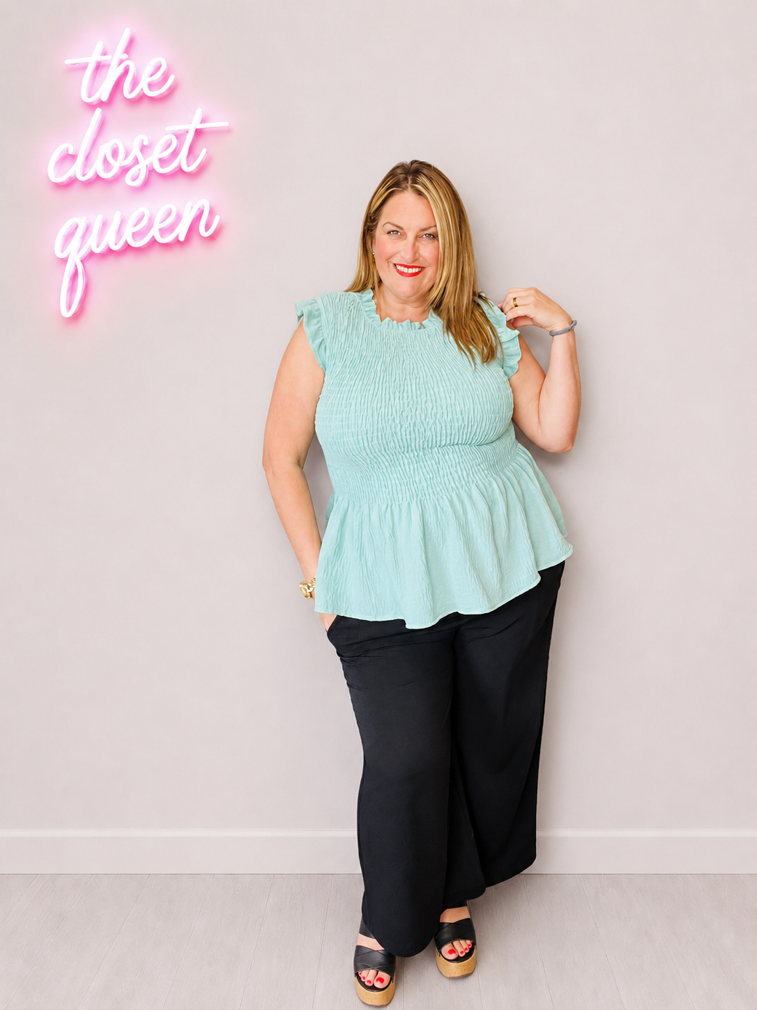 Woman wearing a light blue top and black pants standing in front of a neon sign on a white wall.