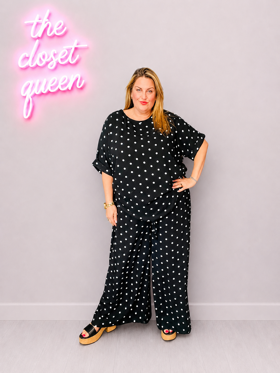 Woman wearing a black polka dot co ord standing in front of a neon sign that reads 'the closet queen' on a gray background.