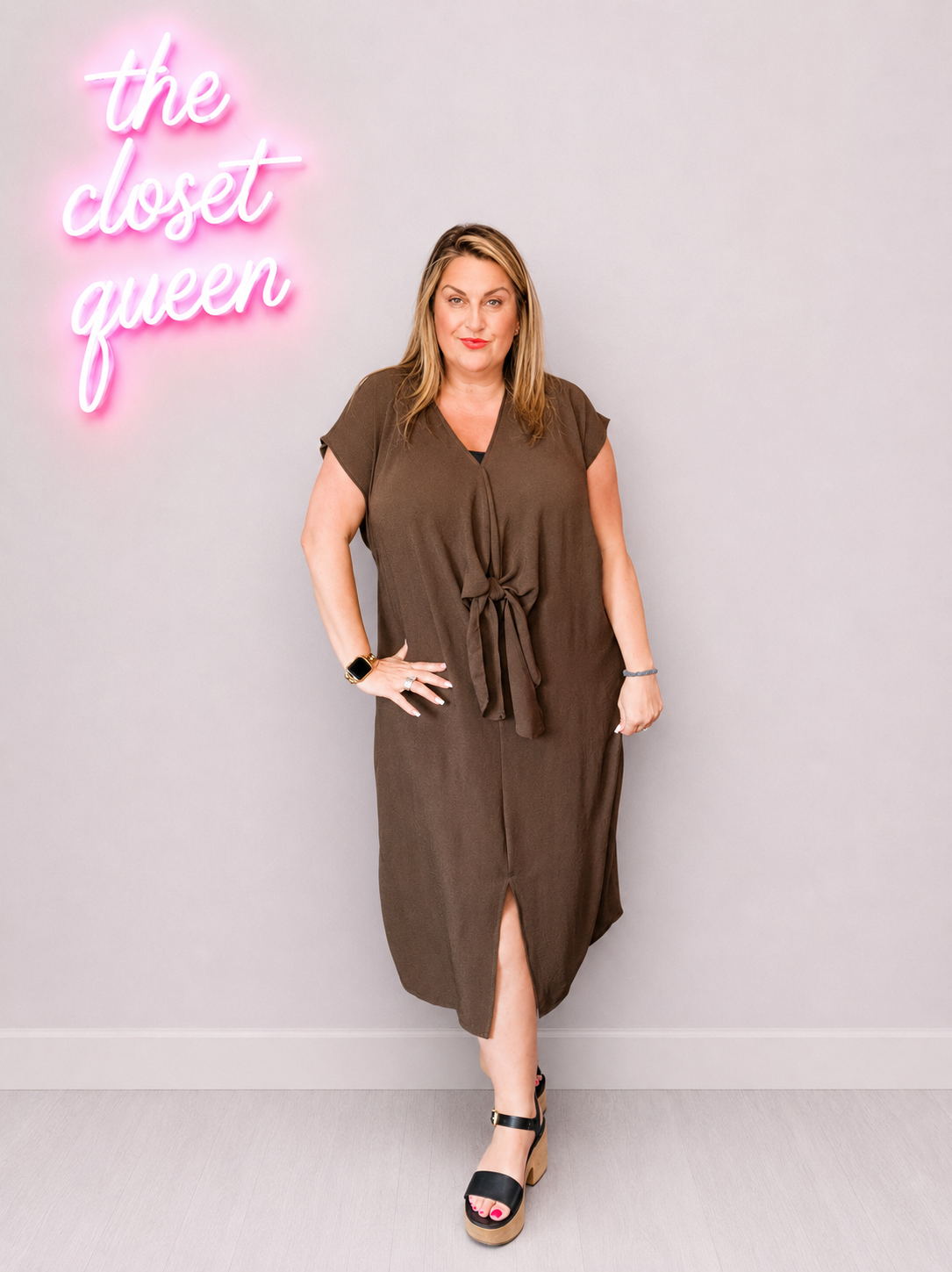 Woman wearing a brown dress with a neon sign reading 'the closet queen' in the background.
